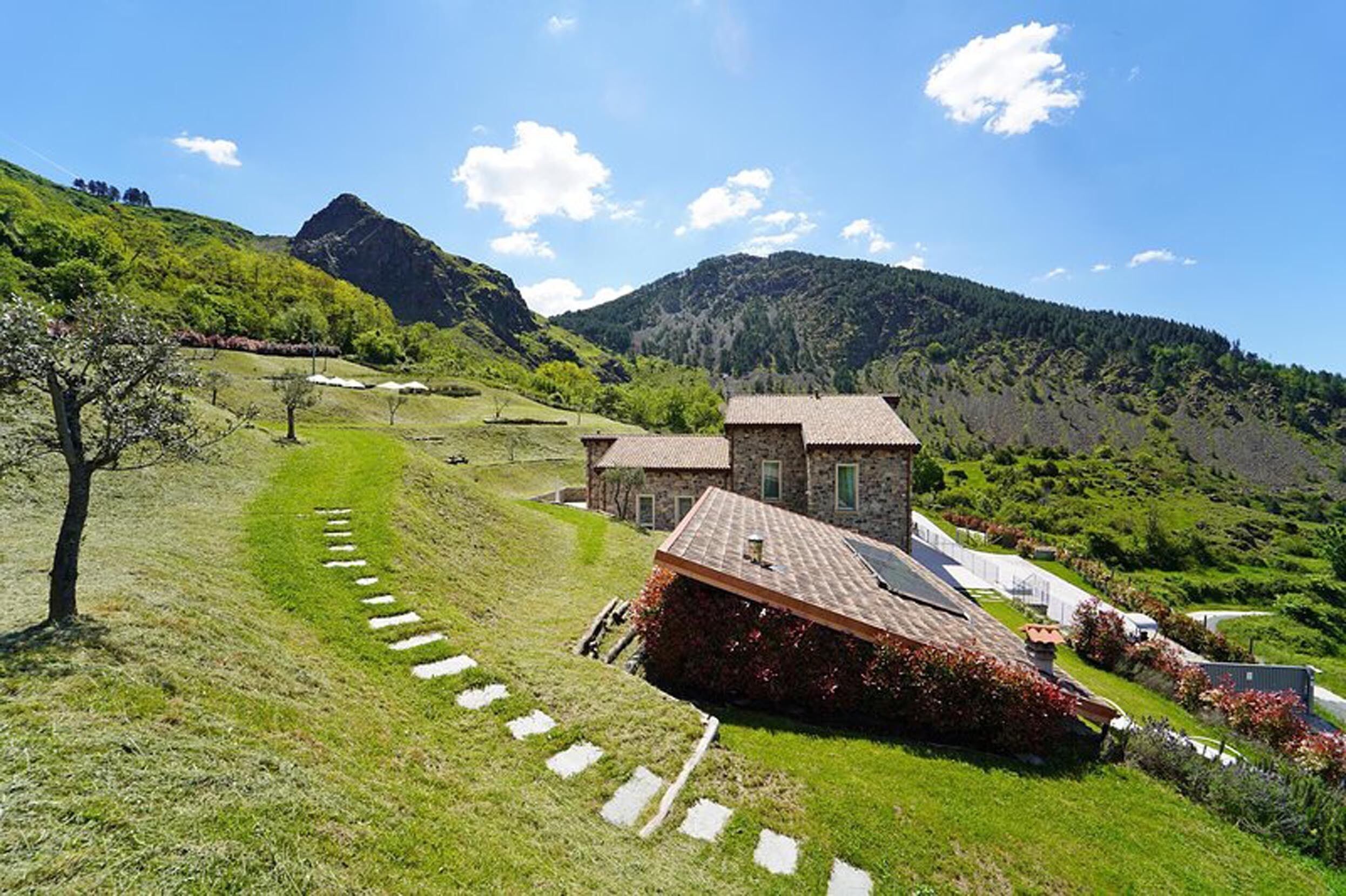 Una casa in pietra con tetto spiovente, immersa tra dolci colline verdeggianti e un cielo sereno.