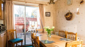 Ein Essbereich mit einem Tisch, Stühlen und einer Blumenvase. Es gibt ein Fenster mit Vorhängen, eine Wanduhr und ein gerahmtes Bild.