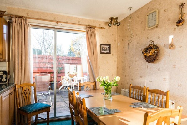 A dining area with a table, chairs, and a vase of flowers. There is a window with curtains, a wall clock, and a framed picture. - Ardgarry Holiday House (Inverness)