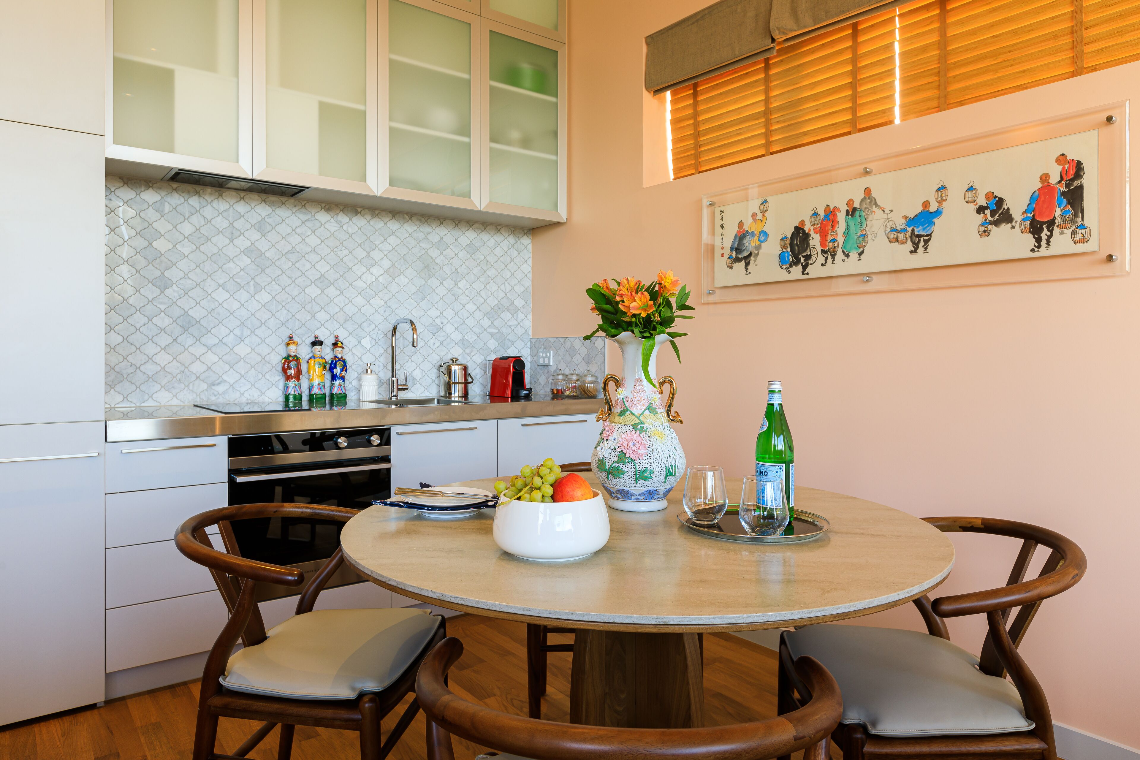 Uma cozinha com armários brancos, painel de azulejos, uma mesa de jantar redonda com cadeiras, um vaso com flores e um quadro na parede.