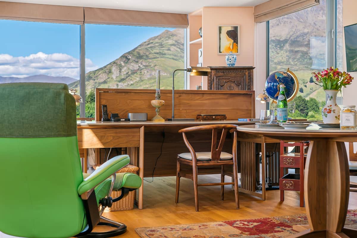 A room with a wooden desk, a green chair, a dining table, and a view of mountains through large windows.