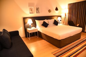 A hotel room with a large bed, two bedside tables with lamps, a black sofa, a striped rug, and a view of the city through the window. - The Port Hotel (Visakhapatnam)
