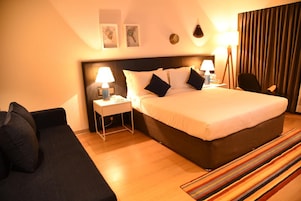 Un cuarto de hotel con una cama grande, dos mesitas de noche con lámparas, un sofá negro, un tapete a rayas y vistas a la ciudad a través de la ventana.
