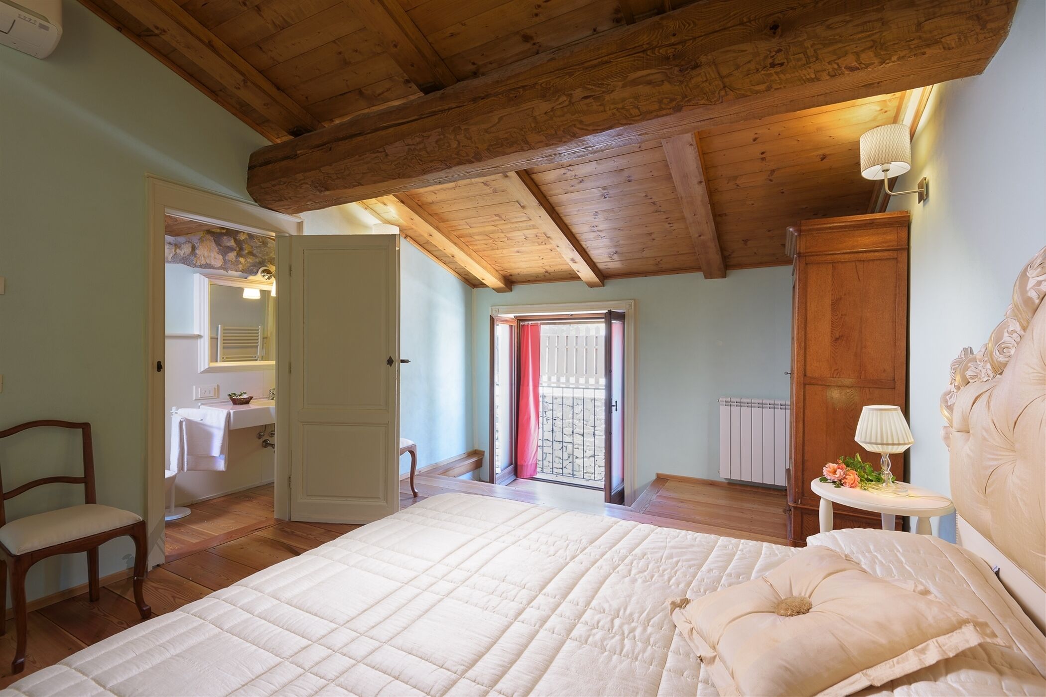 Un dormitorio con techo de madera, una cama con colcha blanca, una mesita de noche con lámpara y vistas a un balcón.