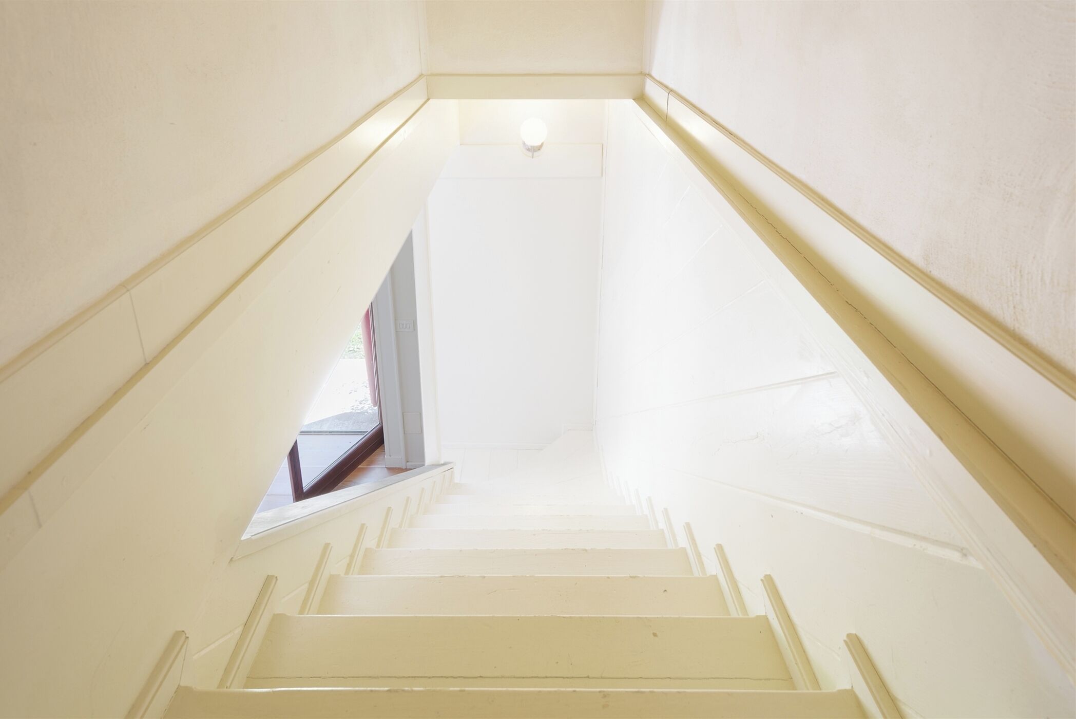 Una escalera blanca y estrecha con tragaluz en la parte superior, que lleva a una puerta.