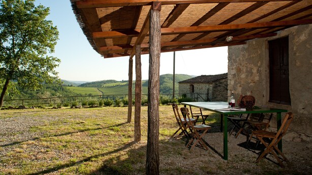 Un espace de restauration extérieur rustique, agrémenté d’une pergola en bois, d’une table avec des chaises et offrant une vue sur des collines verdoyantes en arrière-plan.
