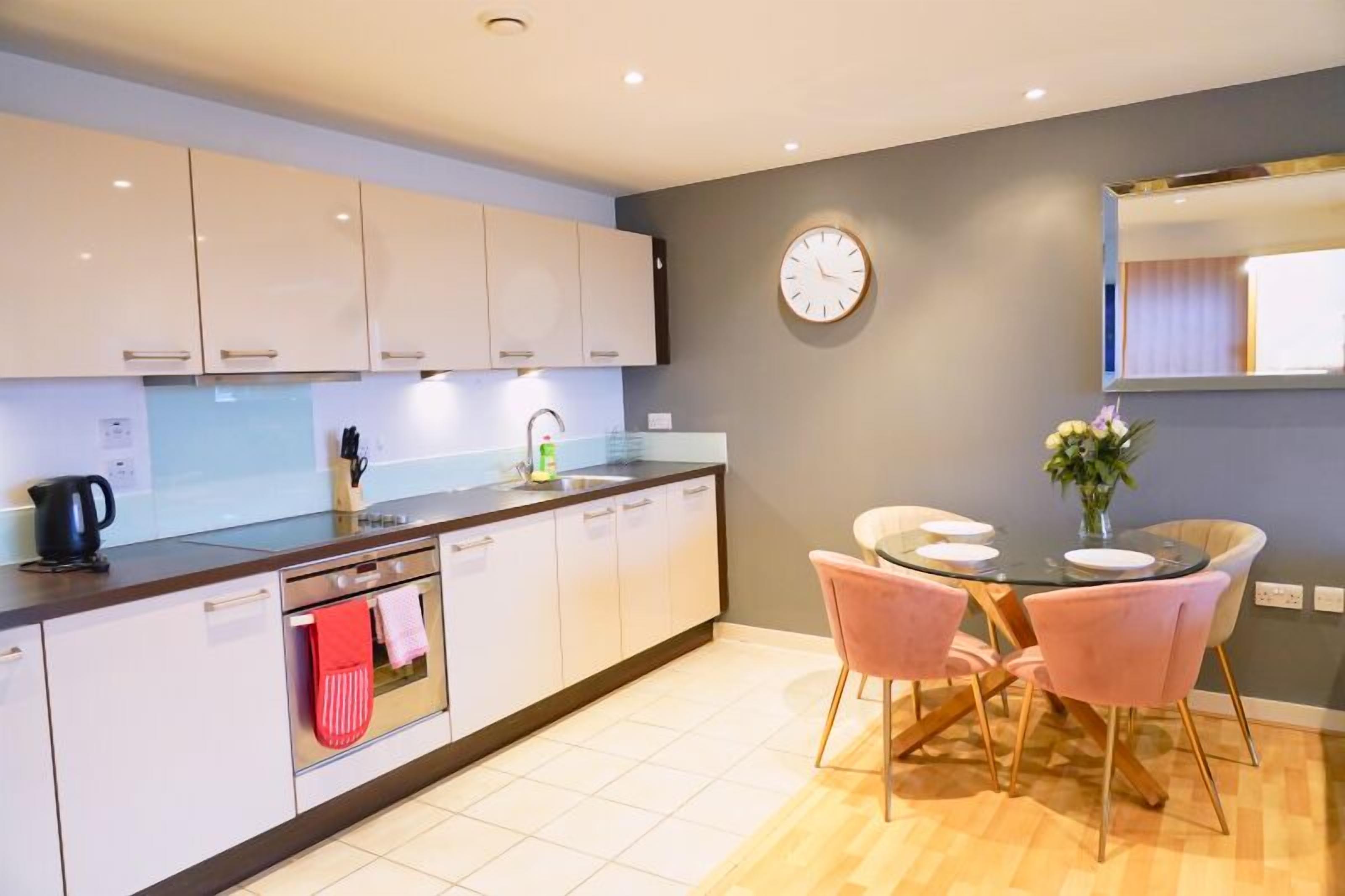 Una cocina moderna con armarios blancos, un hervidor negro, un fregadero y una zona de comedor con una mesa redonda y sillas rosadas.