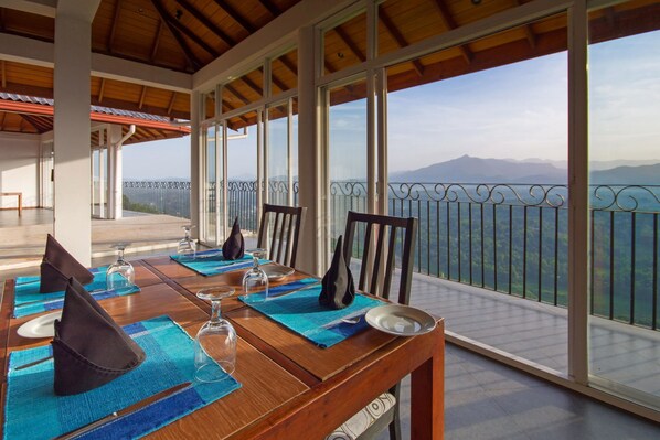 A dining table set with blue placemats, glasses, and napkins on a balcony with a mountain view.