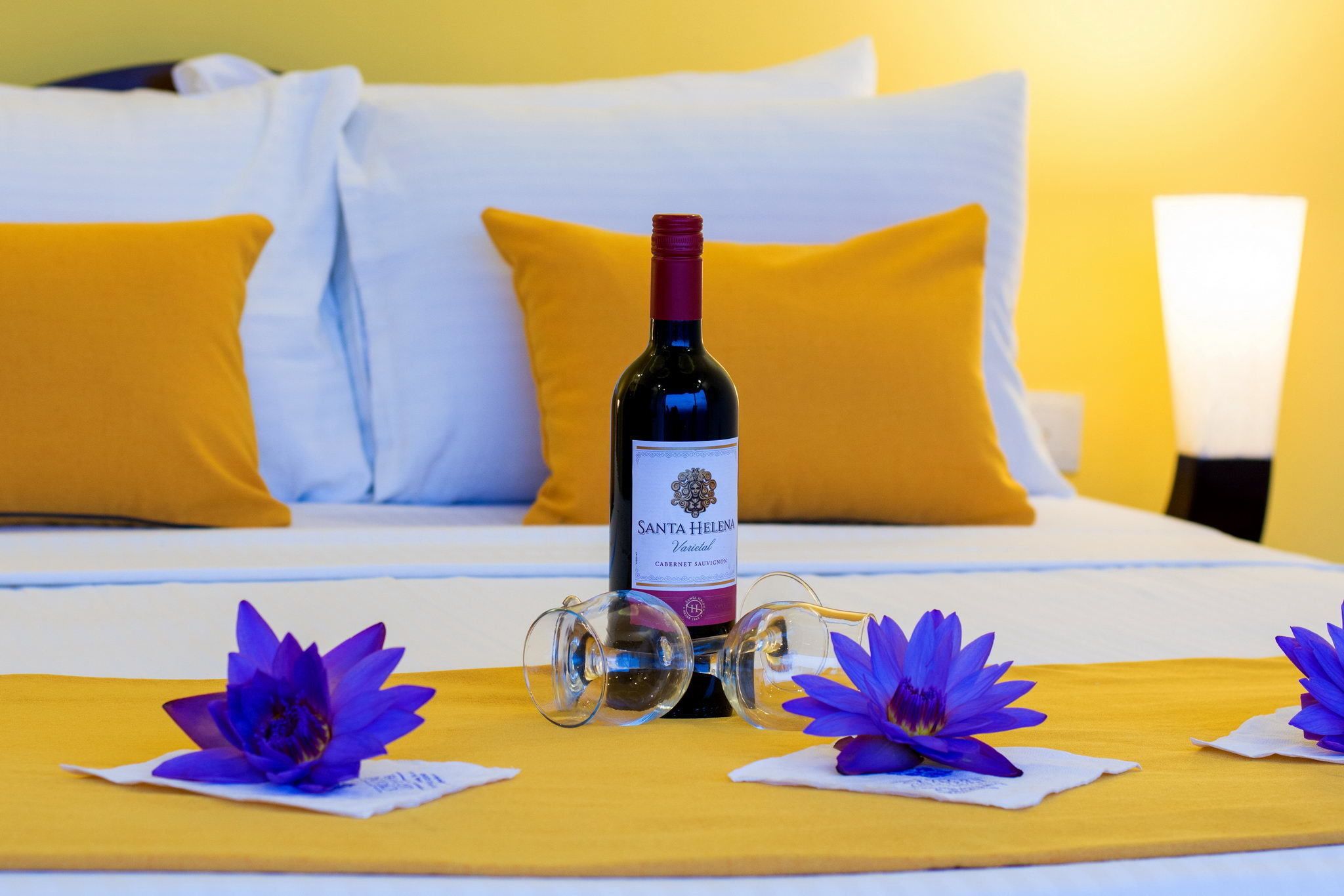 A bed with yellow pillows, a bottle of Santa Helena wine, and purple flowers on a yellow bedspread.