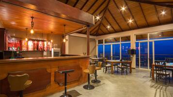 Un restaurante con barra de madera, taburetes y mesas. Amplias ventanas ofrecen vistas de la ciudad de noche.