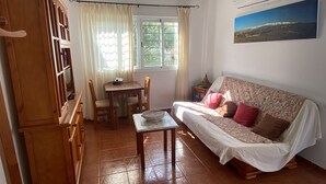 A cozy living room with a sofa, a dining table with chairs, a wooden cabinet, and a window with curtains. - Rent cozy apartment (Nivar)