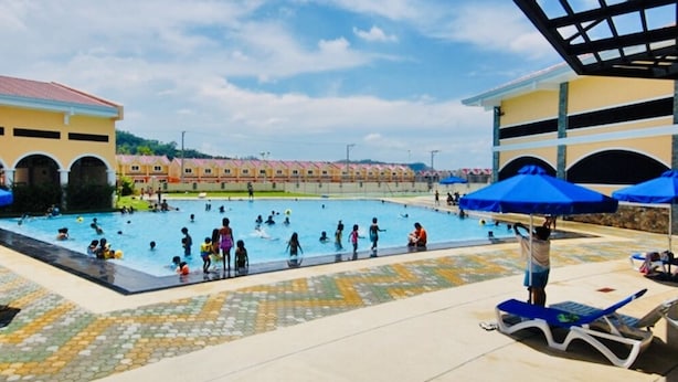 A swimming pool area with multiple people swimming and lounging. There are blue umbrellas and lounge chairs around the pool. - ENTIRE HOUSE/ FULLY AIR CONDITIONED/ HiSpeed Wifi. Cable TV. Smart TV (Pampanga)