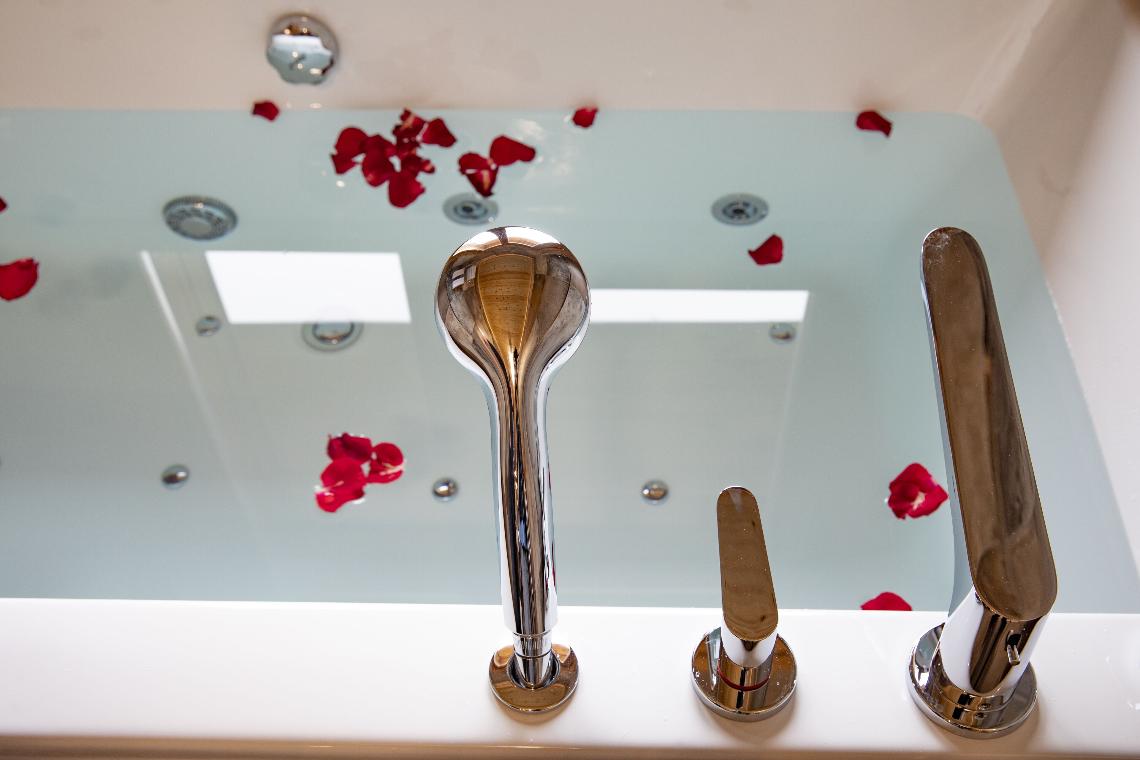 Un baño con bañera de hidromasaje con mampara de vidrio, pétalos de rosa esparcidos sobre la superficie y dos accesorios de cromo.