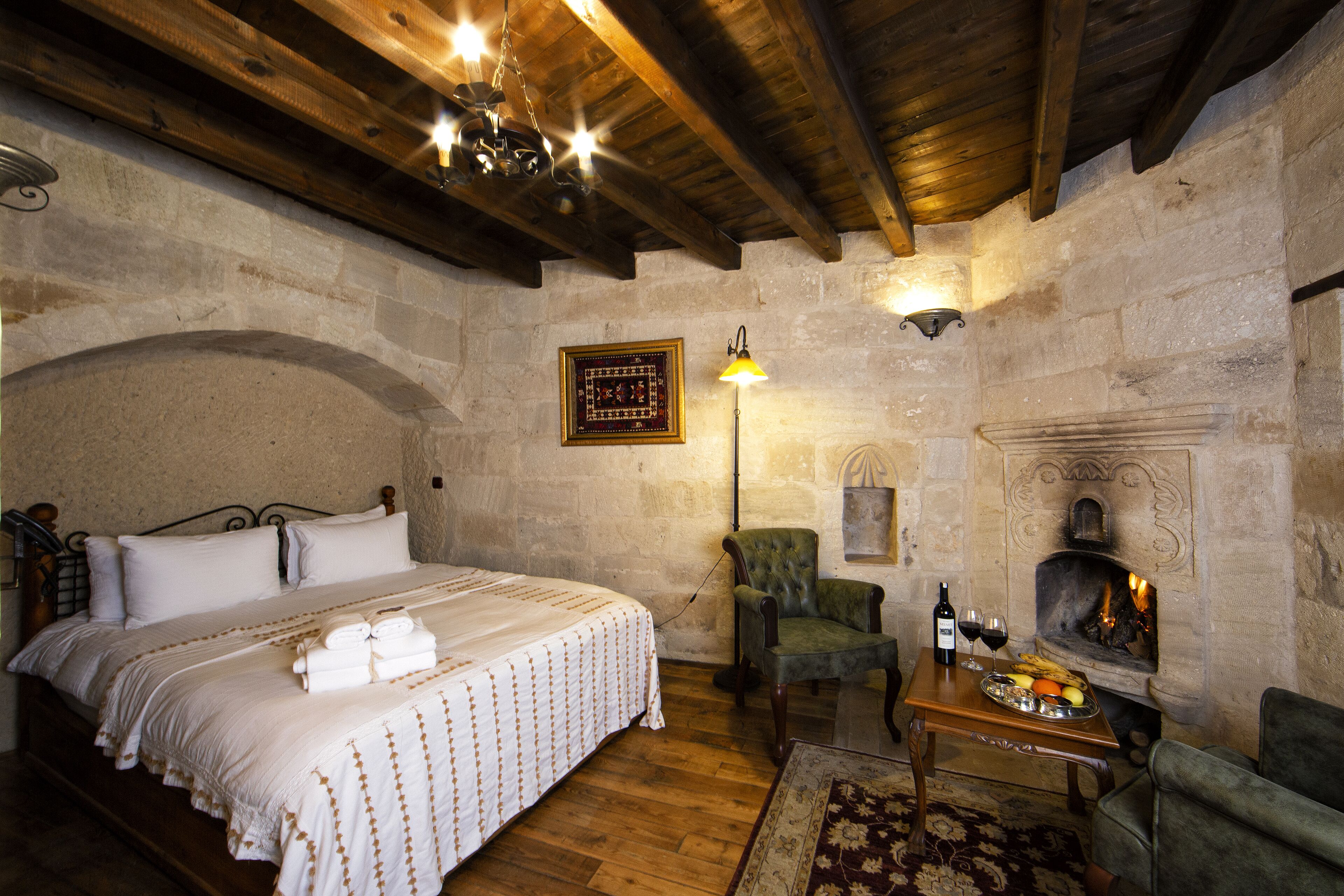 A bedroom with a large bed, a fireplace, a green armchair, and a table with a fruit tray.