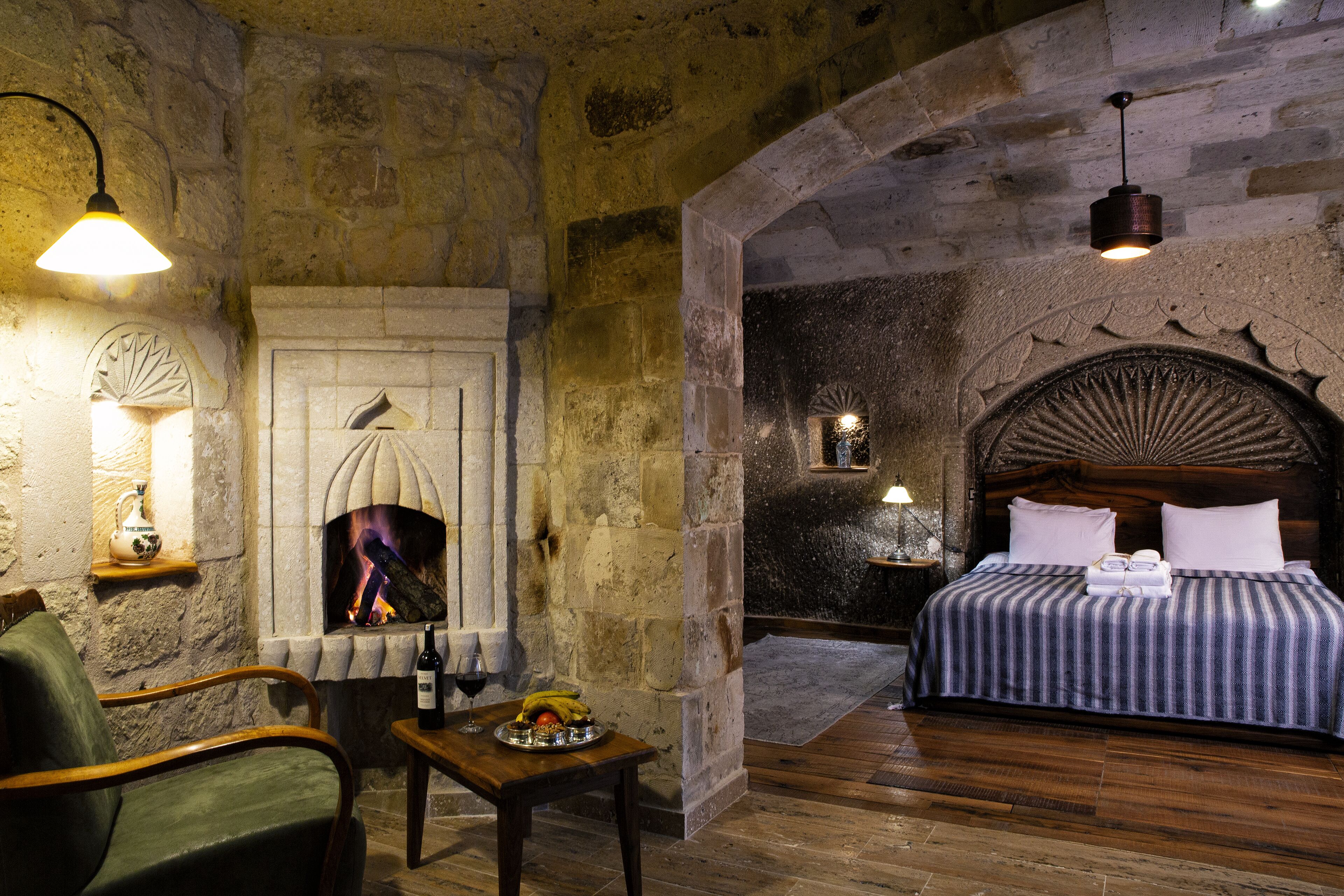 A room with a stone fireplace, a bed with a striped bedspread, a wooden table with a bottle and glasses, and a green armchair.