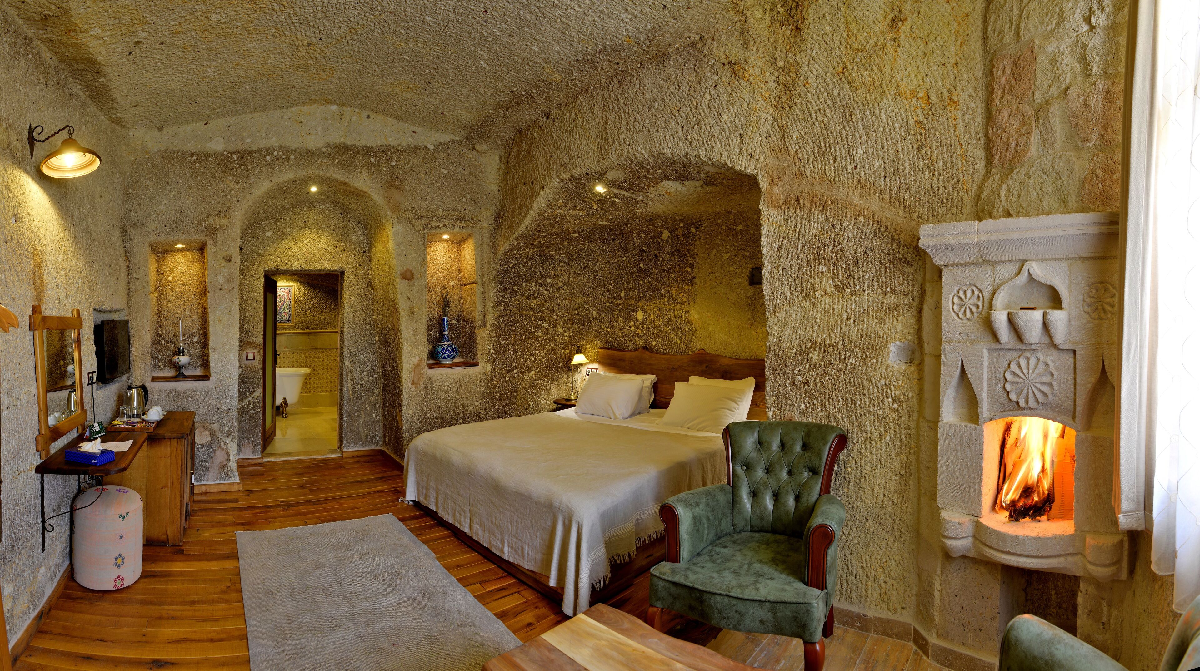 A stone-walled bedroom with a bed, a green armchair, a fireplace, and a wooden floor.
