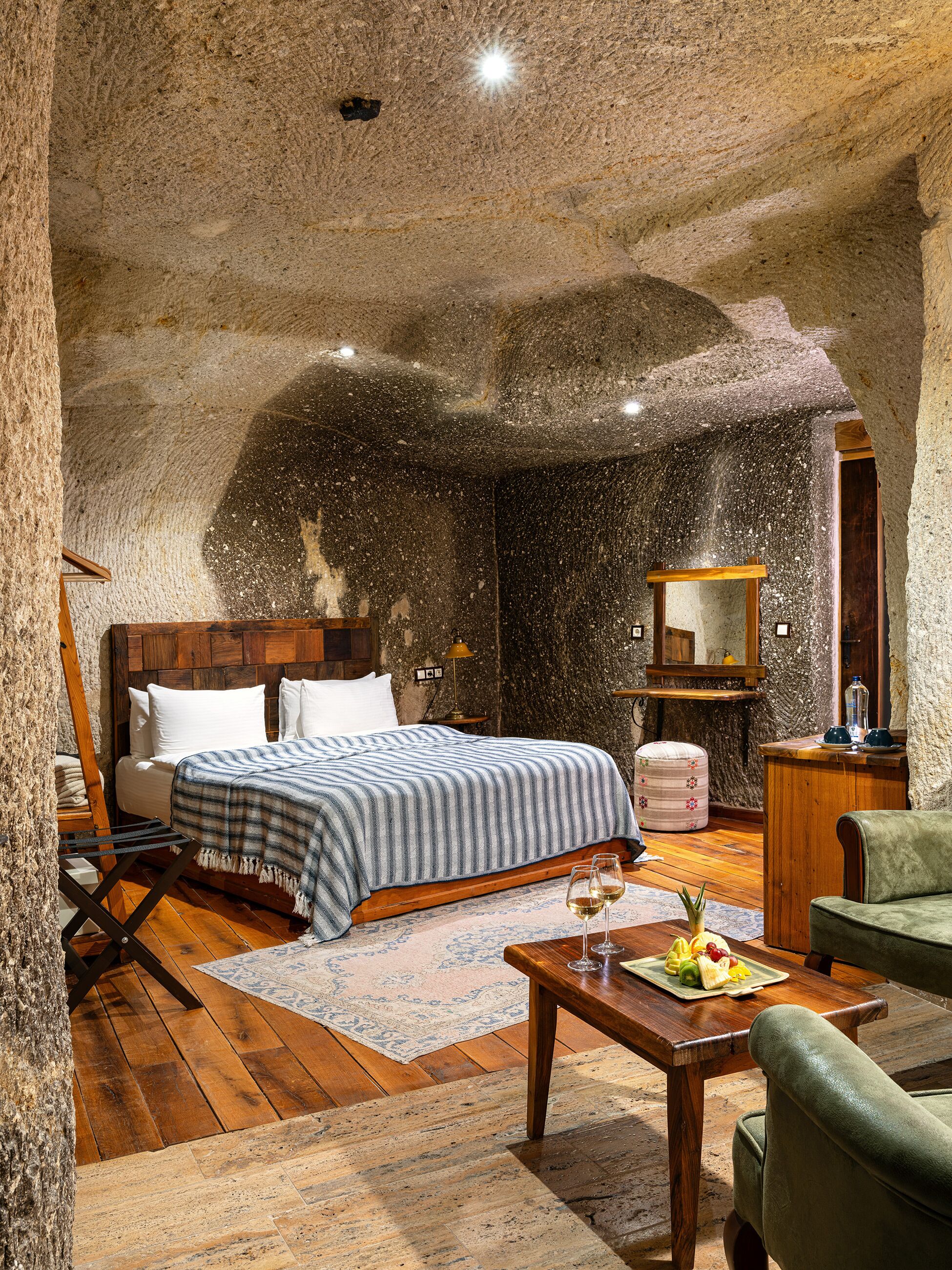 Une chambre à coucher rustique avec un lit en bois, une petite table sur laquelle sont disposés des fruits et des verres à vin, un fauteuil vert et un mur en pierre.