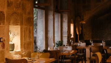 A rustic dining area with wooden tables and chairs, stone walls, and a warm, dimly lit ambiance.