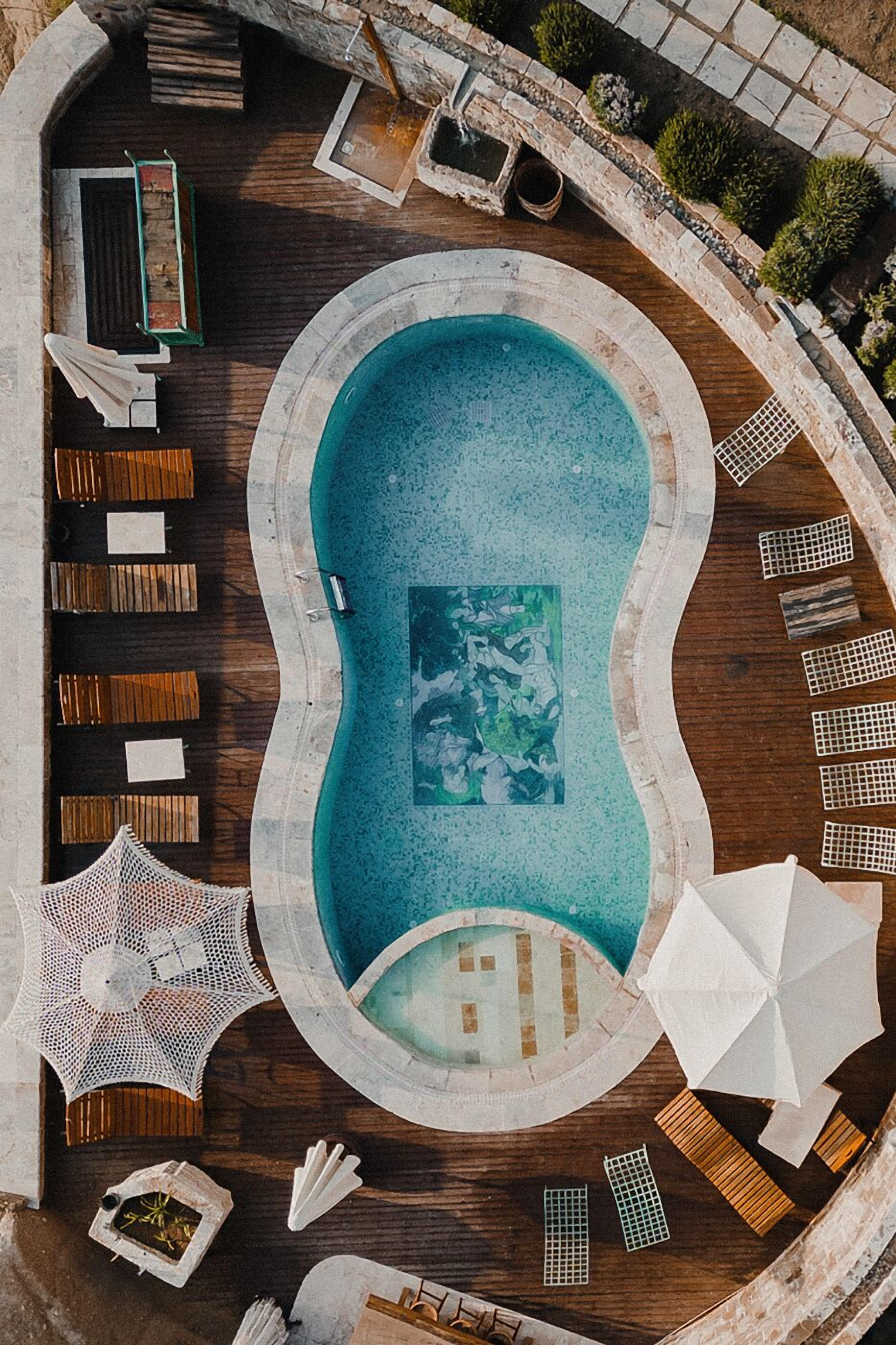 An aerial view of a swimming pool with a wooden deck, lounge chairs, umbrellas, and a mosaic tile border.