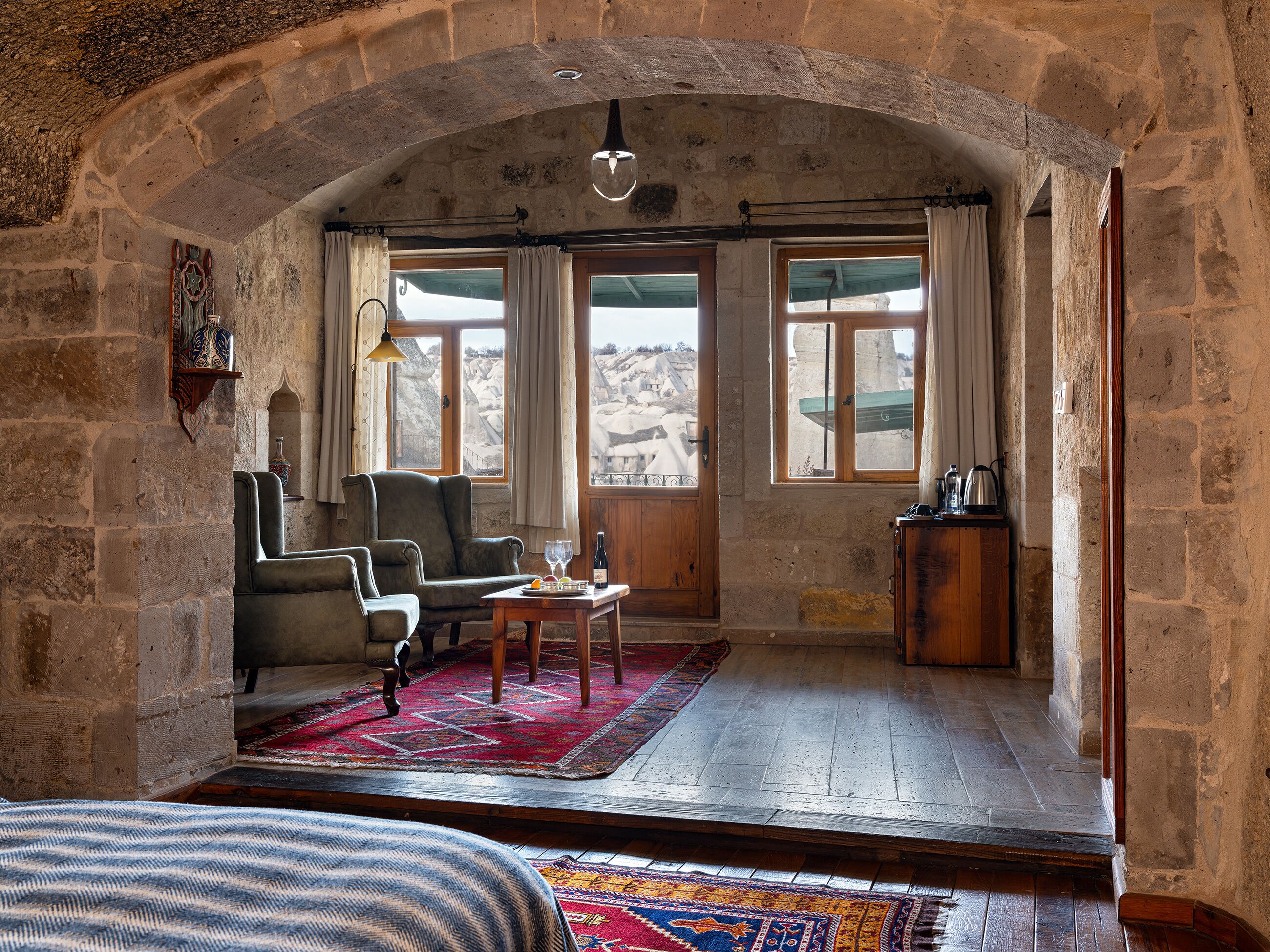 A rustic room with stone walls, a wooden table, a sofa, and a view of a mountainous landscape through large windows.