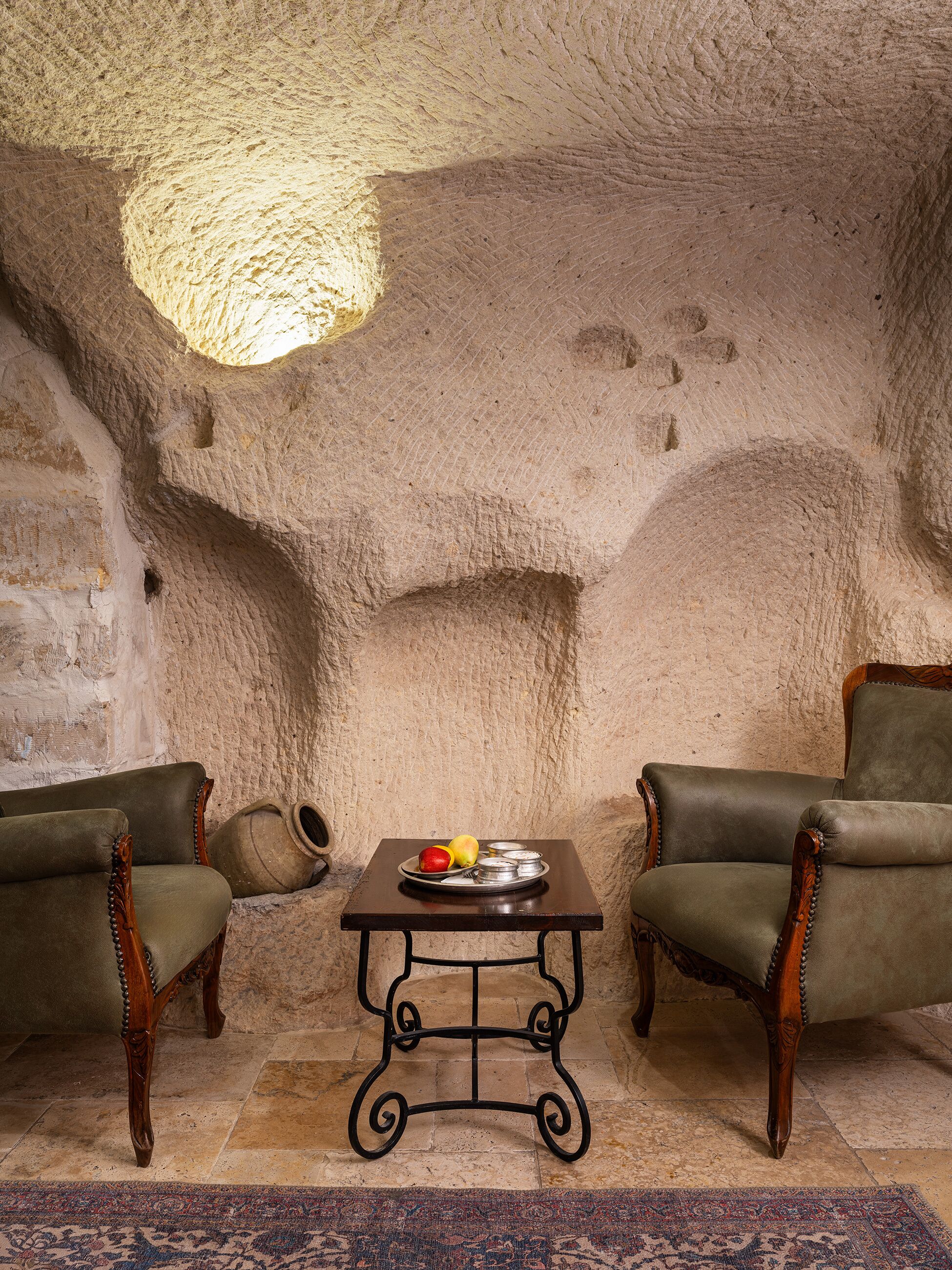 A room with a rustic stone wall, two leather armchairs, a small table with a fruit plate, and a patterned rug.
