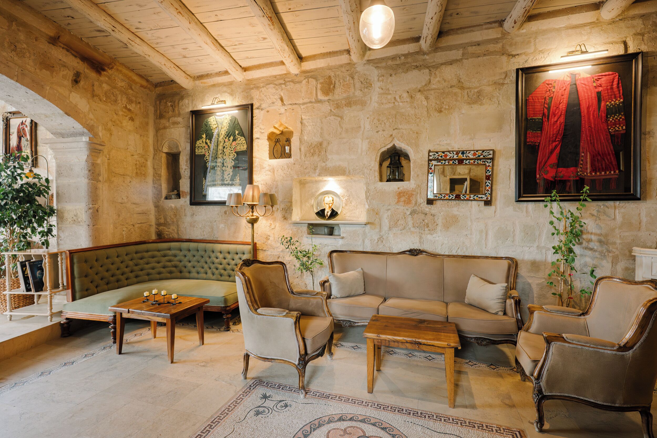 A cozy room with a green sofa, wooden coffee table, and two armchairs. There is a stone wall with a framed tapestry and a mirror.