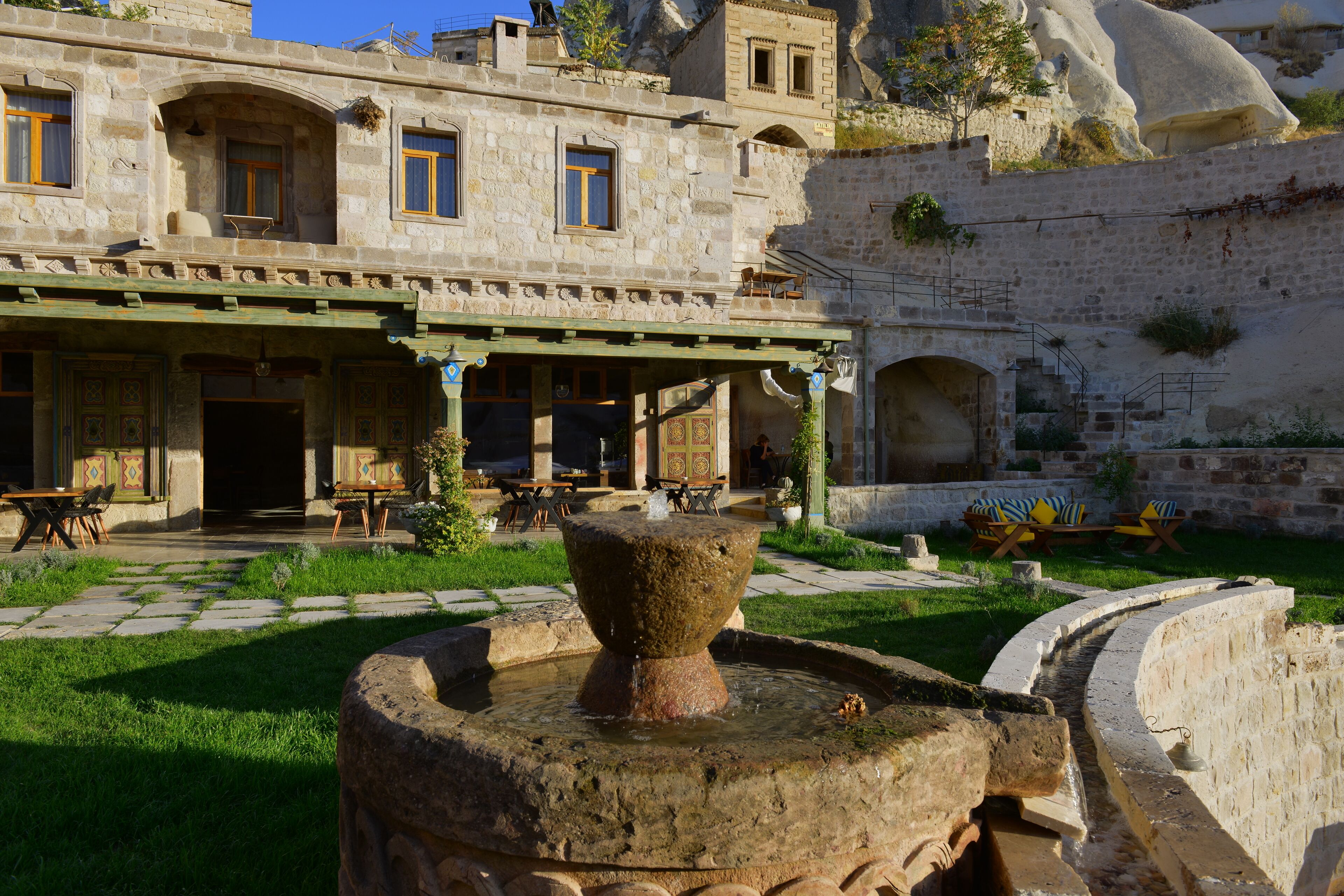 Une cour avec une fontaine en pierre, des sièges en plein air et un bâtiment doté de fenêtres en arc de cercle et d’un patio couvert.