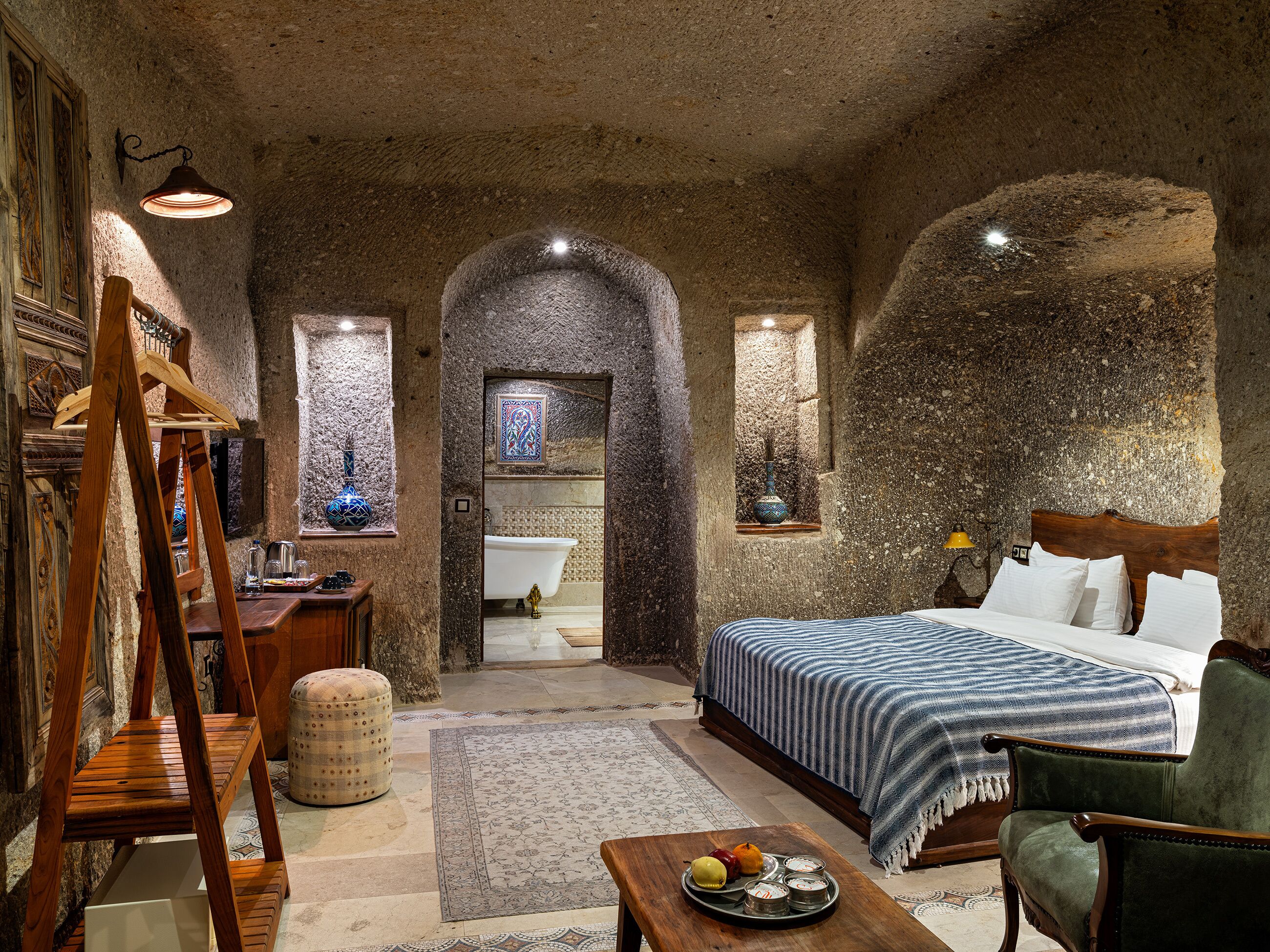 Una habitación rústica con una cama de madera, un sillón verde, una escalera de madera, una pequeña mesa con fruta y vistas a un baño a través de un arco.