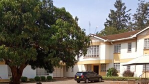Un edificio con entrada para vehículos, un coche aparcado y un jardín ajardinado con flores amarillas y un árbol.