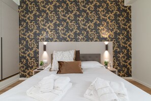 Een net opgemaakt bed met witte lakens en een bruin kussen, met aan weerszijden twee nachtkastjes met lampen, in een kamer met behang met een patroon.