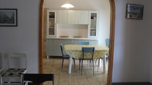 Un comedor con una mesa preparada para cuatro personas, una cocina con armarios blancos y un cuadro en la pared.