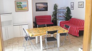 Una habitación pequeña y bien iluminada con una mesa de comedor, sillas, un sofá rojo, un sofá rojo a rayas, un armario blanco y una estantería con objetos decorativos.
