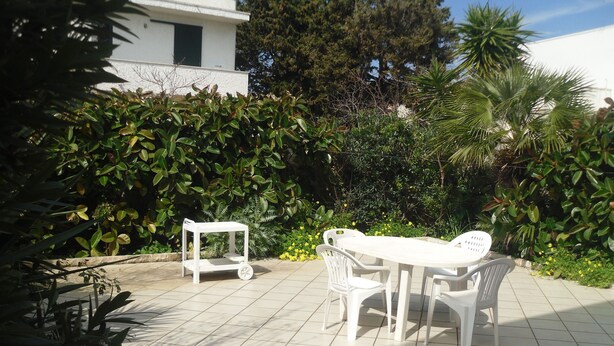 Un patio con una mesa y sillas blancas, rodeado de follaje y un edificio blanco al fondo.