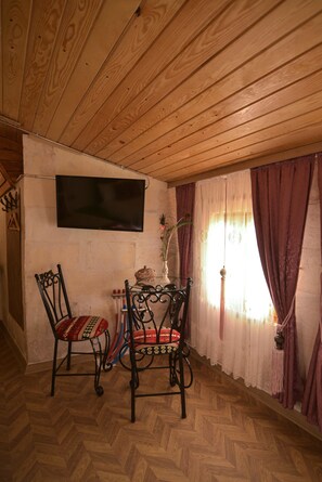 A room with wooden ceiling and walls, a flat-screen TV mounted on the wall, two ornate chairs with patterned cushions, a small table with a vase and decorative items, and a window with purple curtains. - DOĞANBEY KONAĞI (Gaziantep)