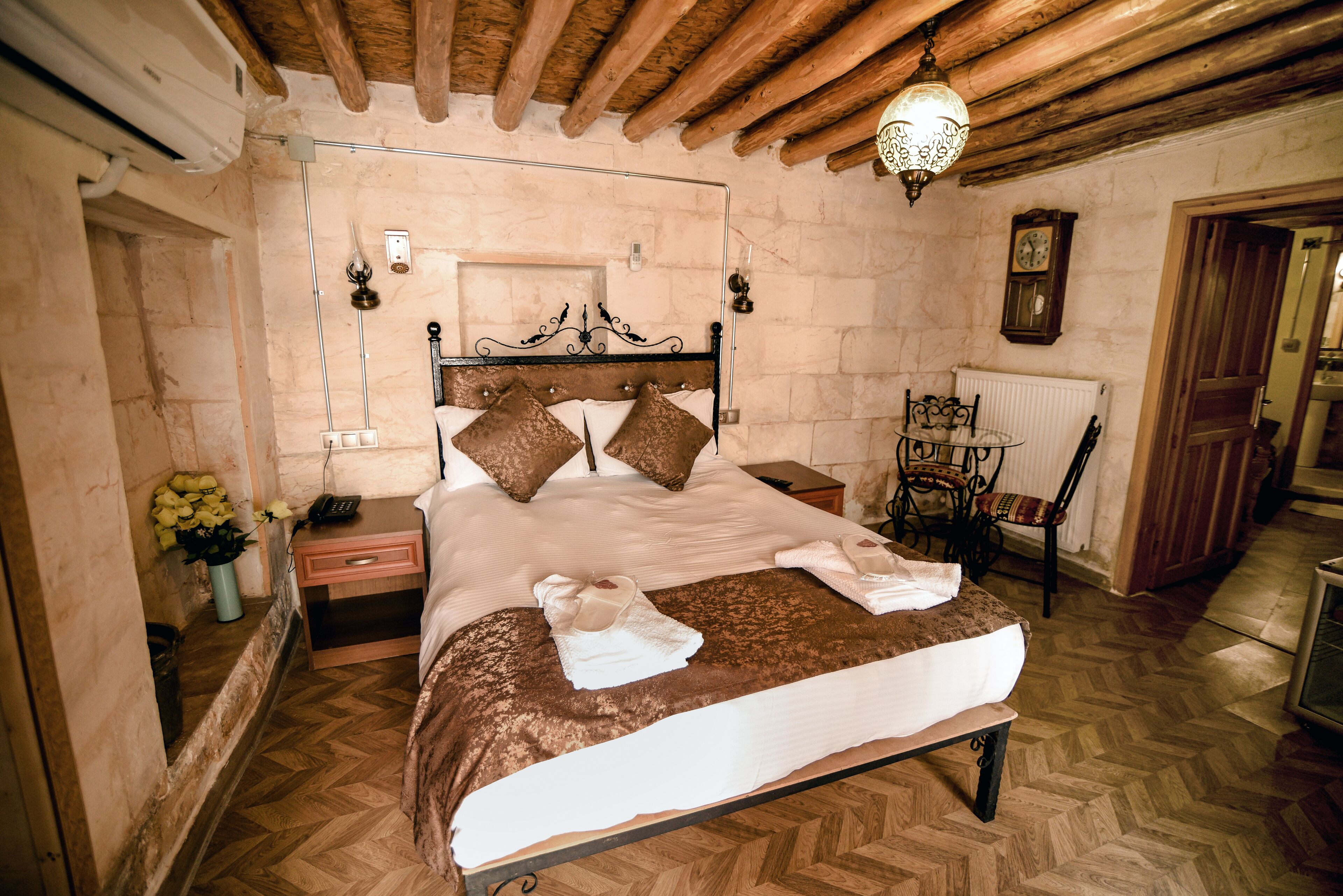Un dormitorio con techo de madera, una cama con ropa de cama marrón, una mesita de noche con lámpara y una silla con cojín.