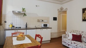 Cocina compacta con armarios blancos, microondas y un lavamanos pequeño. Hay una mesa de comedor con sillas y una cesta de naranjas. Se ve un sofá floral al fondo.