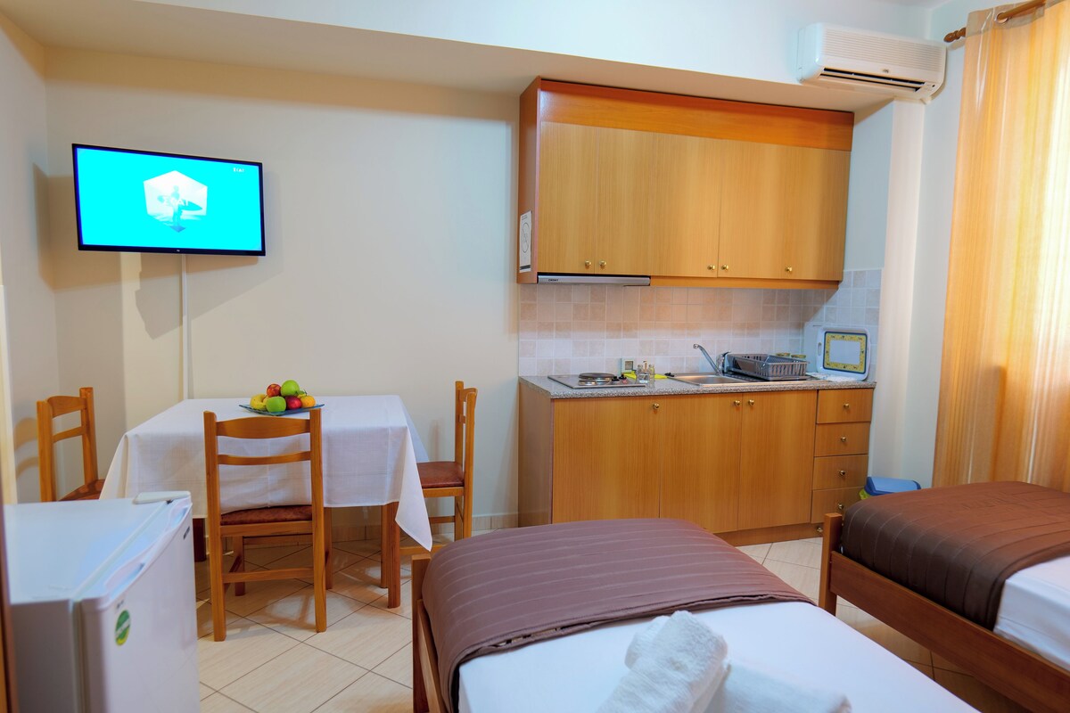 A hotel room with a bed, a desk with a chair, a small kitchenette, and a television mounted on the wall.