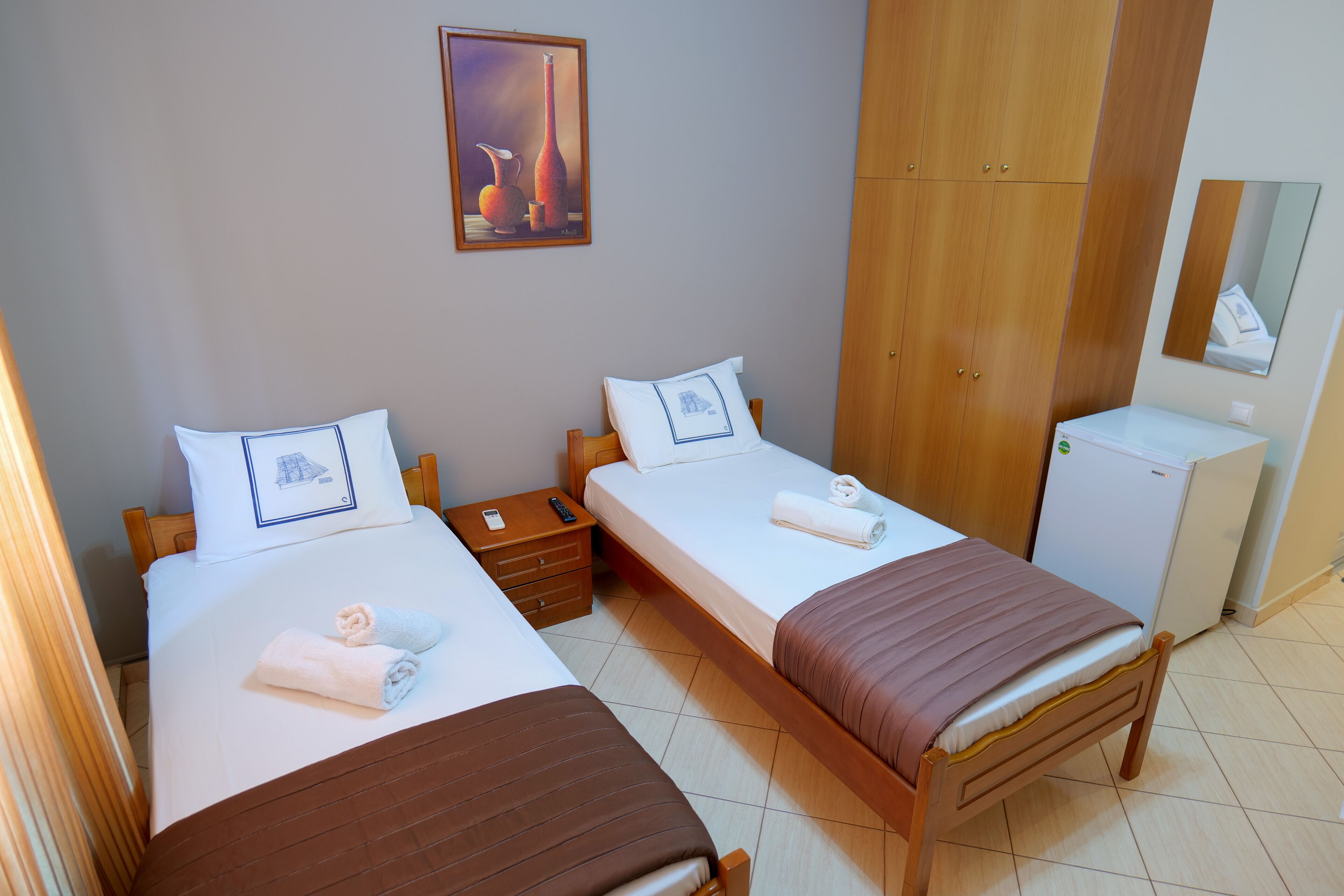 A hotel room with two single beds, a small bedside table with a lamp, a white wall with a framed artwork, a wooden wardrobe, and a small white refrigerator.