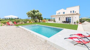 A swimming pool with red lounge chairs, a white building with a patio, and a view of the sea. - Very modern villa with private pool and huge garden in the heart of Mallorca. (Llubi)