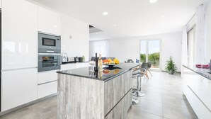 A modern kitchen with white cabinetry, a central island, and a dining area with bar stools. - Very modern villa with private pool and huge garden in the heart of Mallorca. (Llubi)