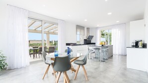 A modern kitchen with a central island, a dining area with a glass table and chairs, and a view of the outdoors. - Very modern villa with private pool and huge garden in the heart of Mallorca. (Llubi)