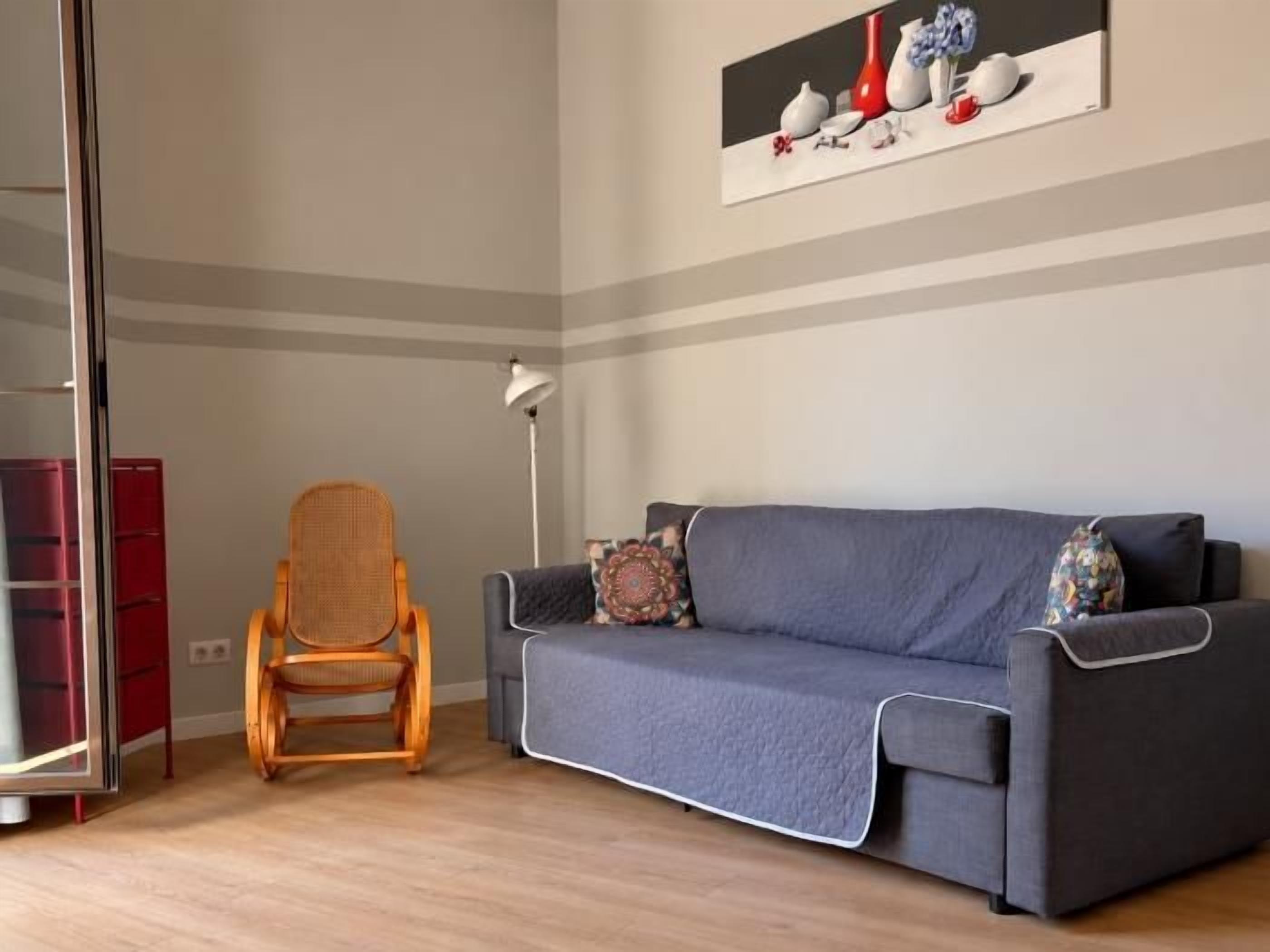 Een kamer met een grijze bank, een rieten stoel, een rode kast en een schilderij aan de muur.