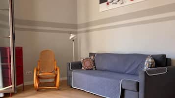 Een kamer met een grijze bank, een rieten stoel, een rode kast en een schilderij aan de muur.
