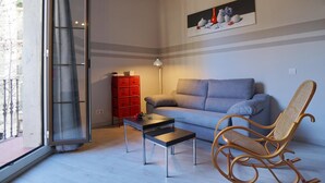 A living room with a grey sofa, a wicker rocking chair, a small table, a red storage unit, and a painting on the wall. - Chic studio in a perfect location in Barcelona (Barcelona)