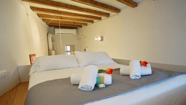 A neatly made bed with white linens and towels, a wooden ceiling, and a small bedside table with a book and a bottle. - Chic studio in a perfect location in Barcelona (Barcelona)