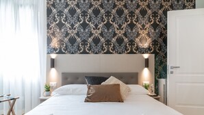 Een netjes opgemaakt bed met witte lakens en een bruin kussen, een nachtkastje met een lamp, en een muur met behang.