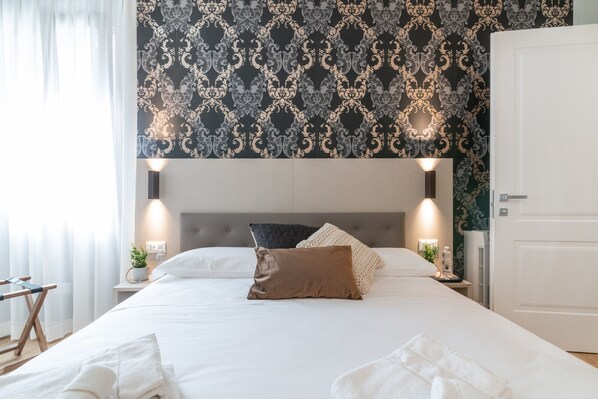 A neatly made bed with white linens and a brown pillow, a bedside table with a lamp, and a wall with a patterned wallpaper.