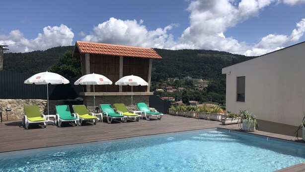 A swimming pool with lounge chairs and umbrellas, surrounded by a wooden deck and a building with a tiled roof. - House of Quinteiro de Azagães (Azagães)