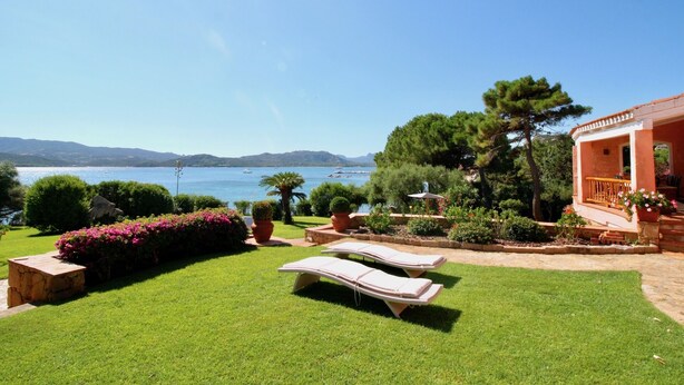 A well-maintained garden with a pool area, including lounge chairs and a table, overlooking a scenic body of water with boats.