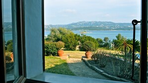 A view from a window overlooking a garden with a pathway, potted plants, and a body of water with distant hills.